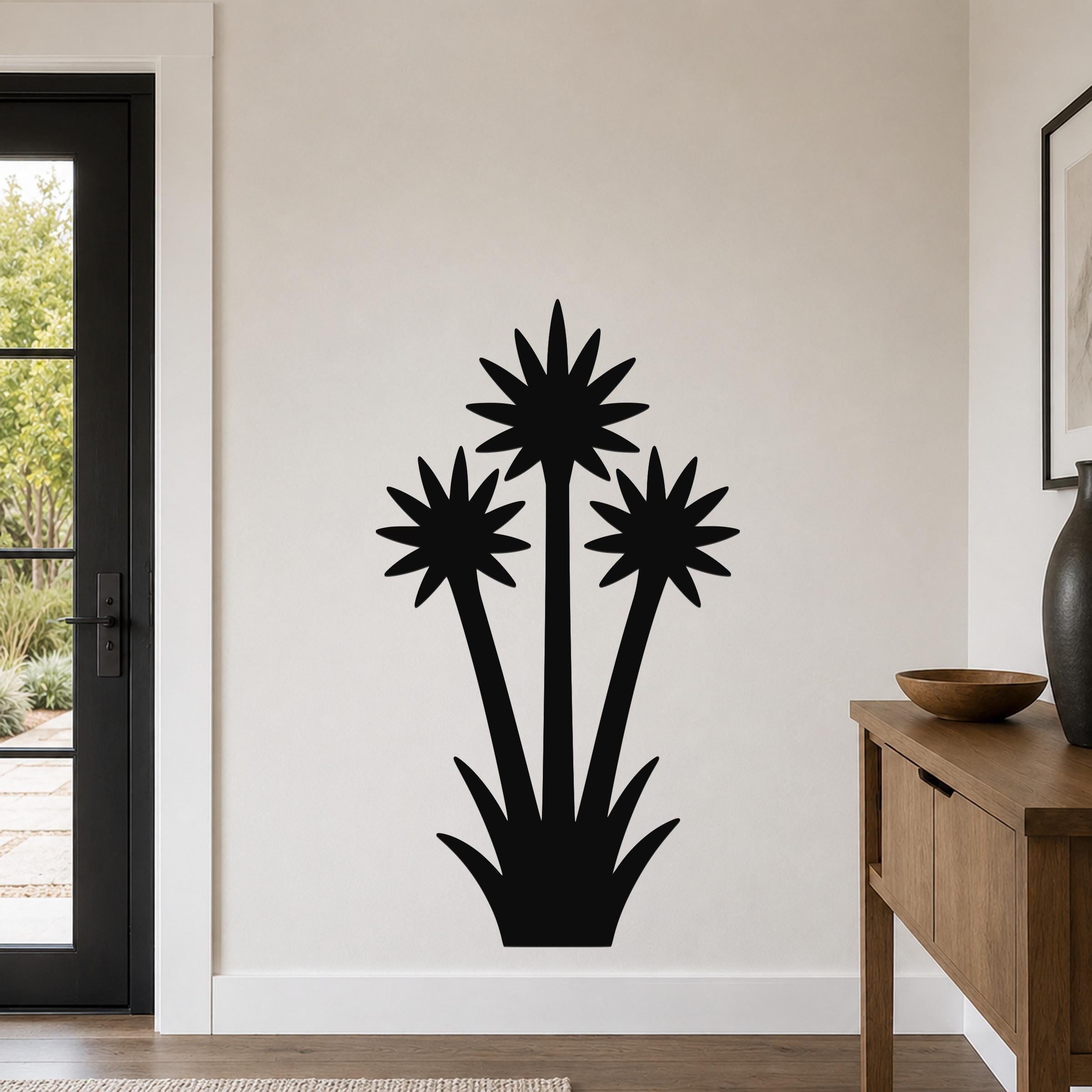 Black metal wall art cutout of a blooming yucca plant with three starburst flowers and spiky base, mounted on a white wall in a living room near a door and console table.