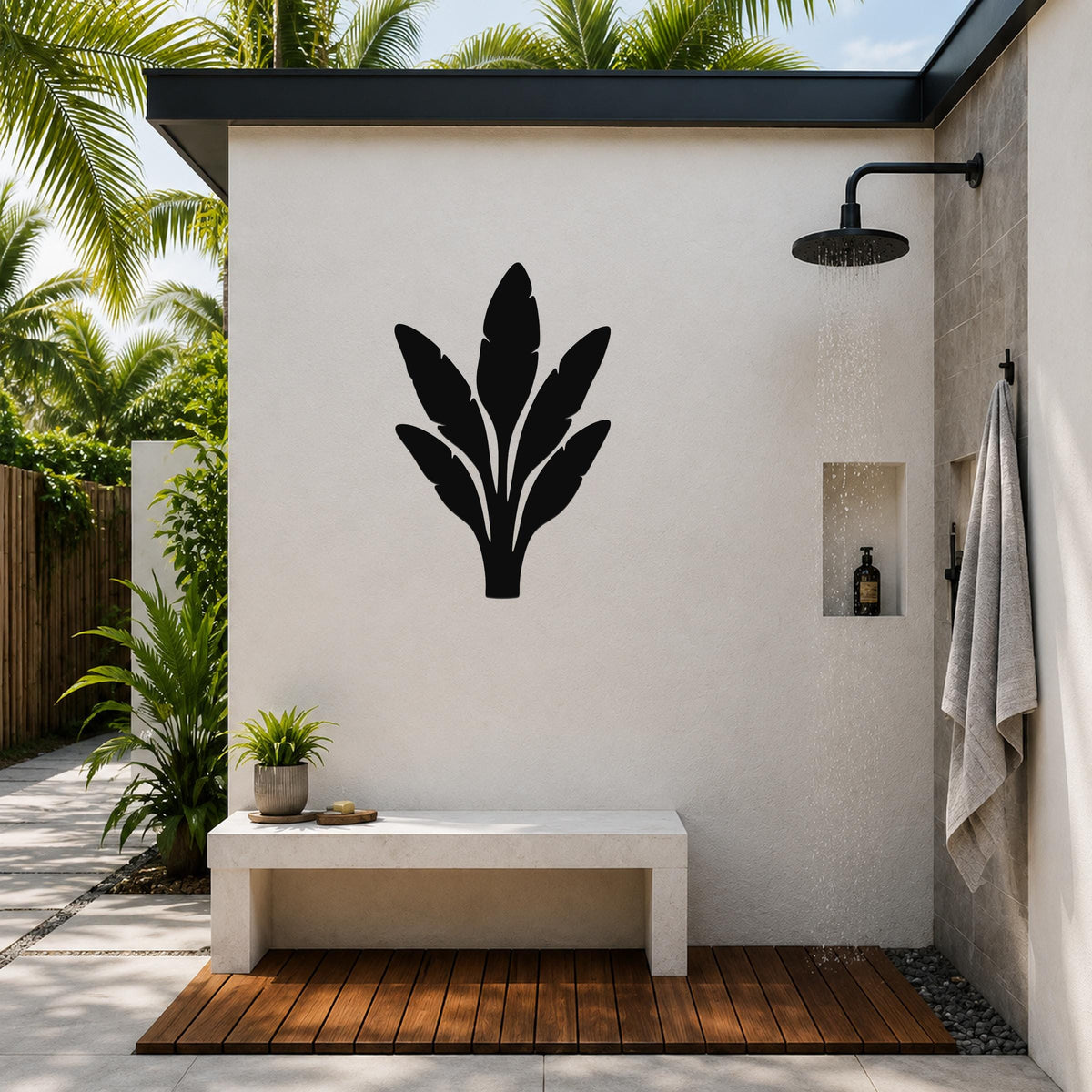 Black metal banana plant wall art silhouette mounted on a white outdoor shower wall, above a concrete bench in a tropical patio setting.