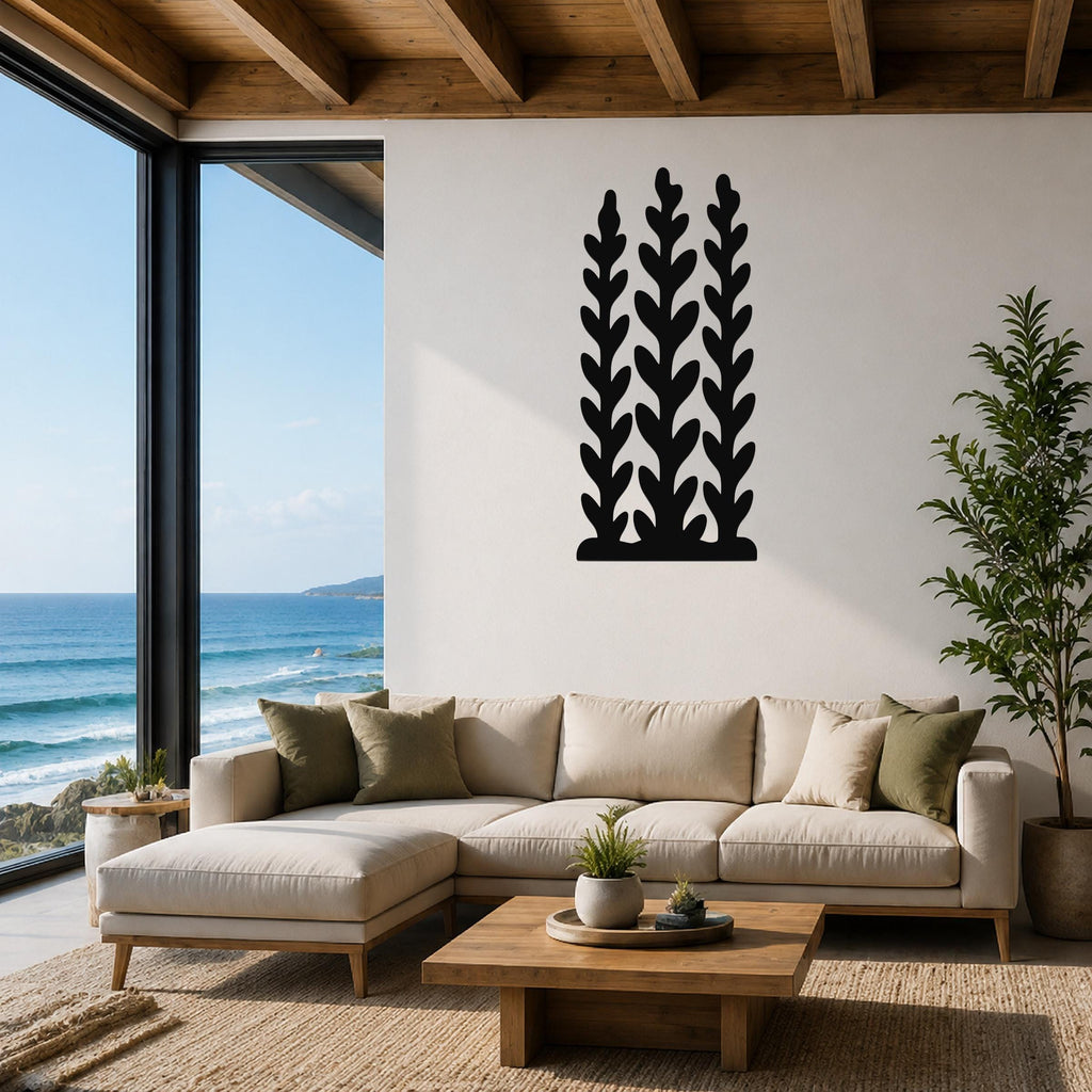 Black metal wall art with a kelp and seaweed silhouette design, mounted on a white wall above a beige sectional sofa in a living room with ocean views.
