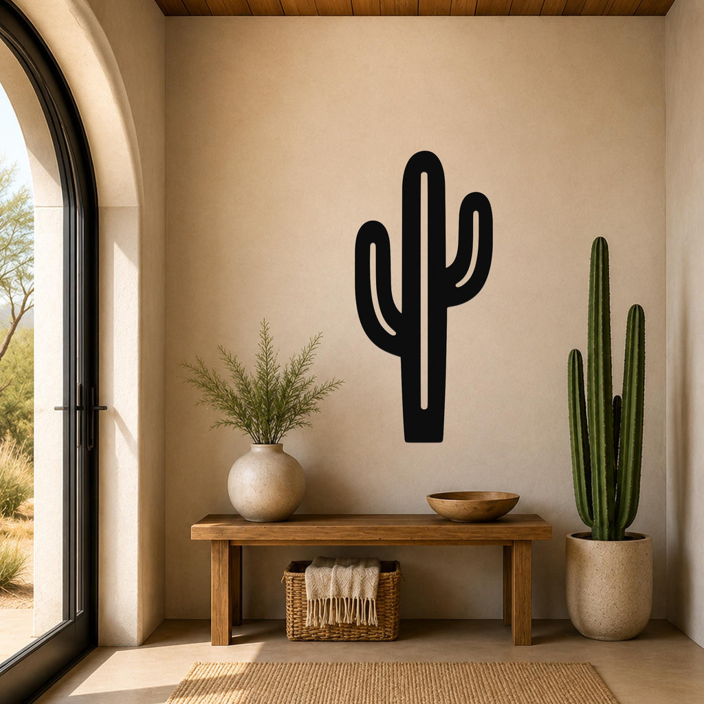 Black metal saguaro cactus wall art with cutout branches, mounted on a beige wall above a wooden bench in a sunlit entryway.