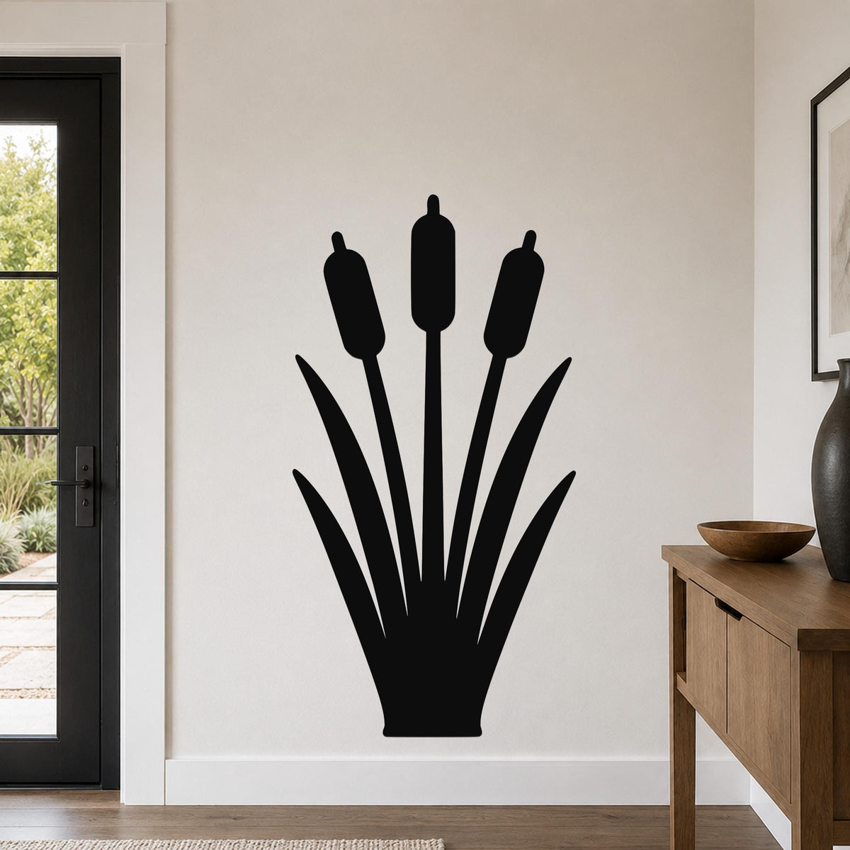 Black metal cattail wall art silhouette with three seed heads and long leaves, mounted on a light-colored wall in a living room near a door and console table.