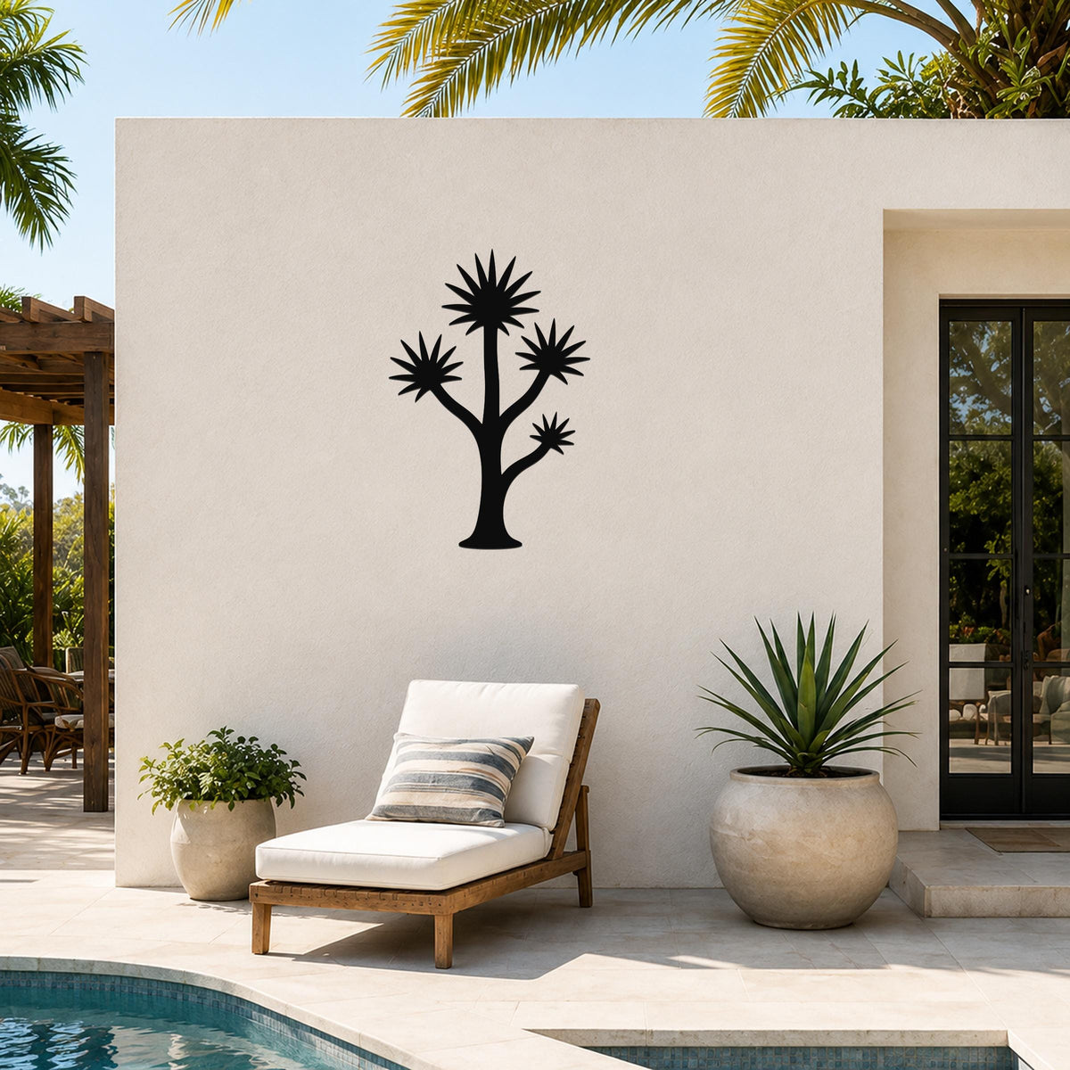 Black metal wall art in a yucca tree silhouette with spiky leaf cutouts, mounted on an outdoor patio wall above a lounge chair.