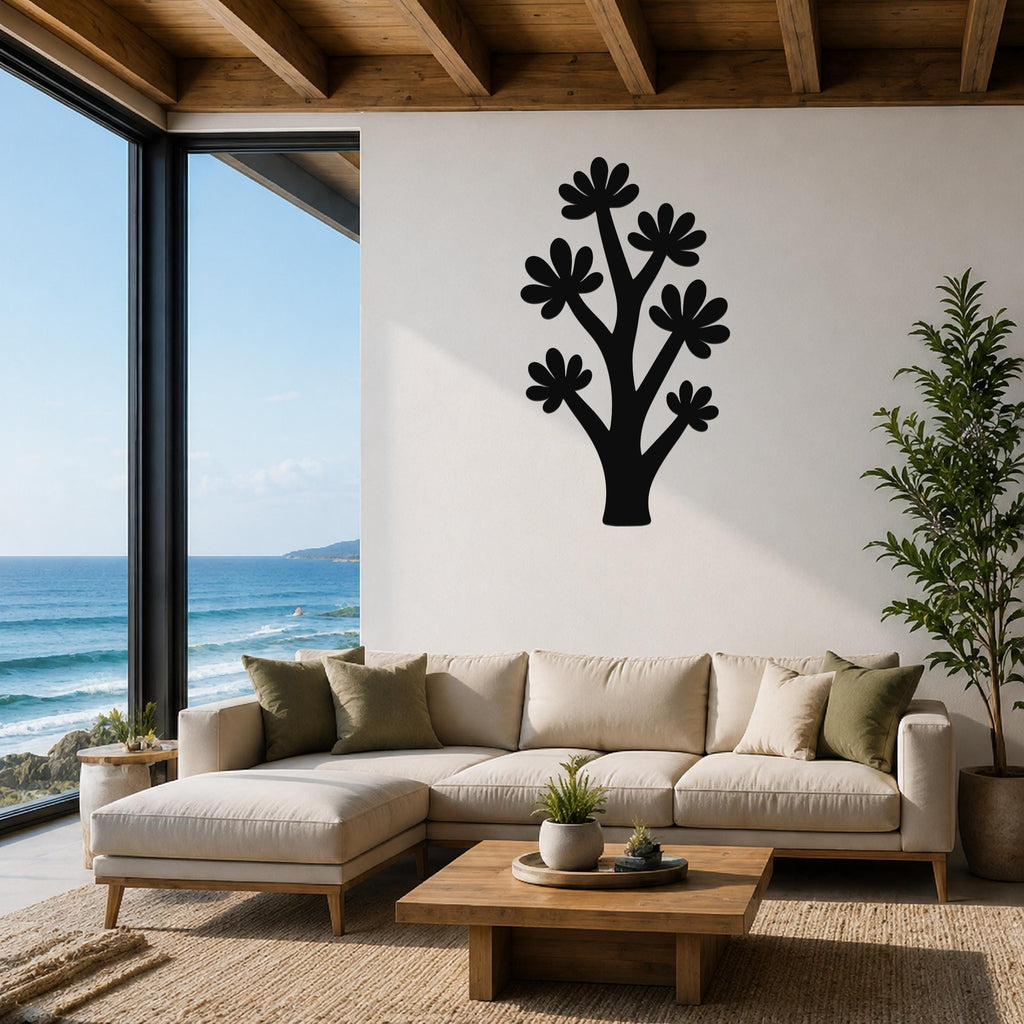 Black metal wall art in a stylized tree shape with flower cutouts, mounted on a white wall above a beige sectional sofa in a living room with ocean views.