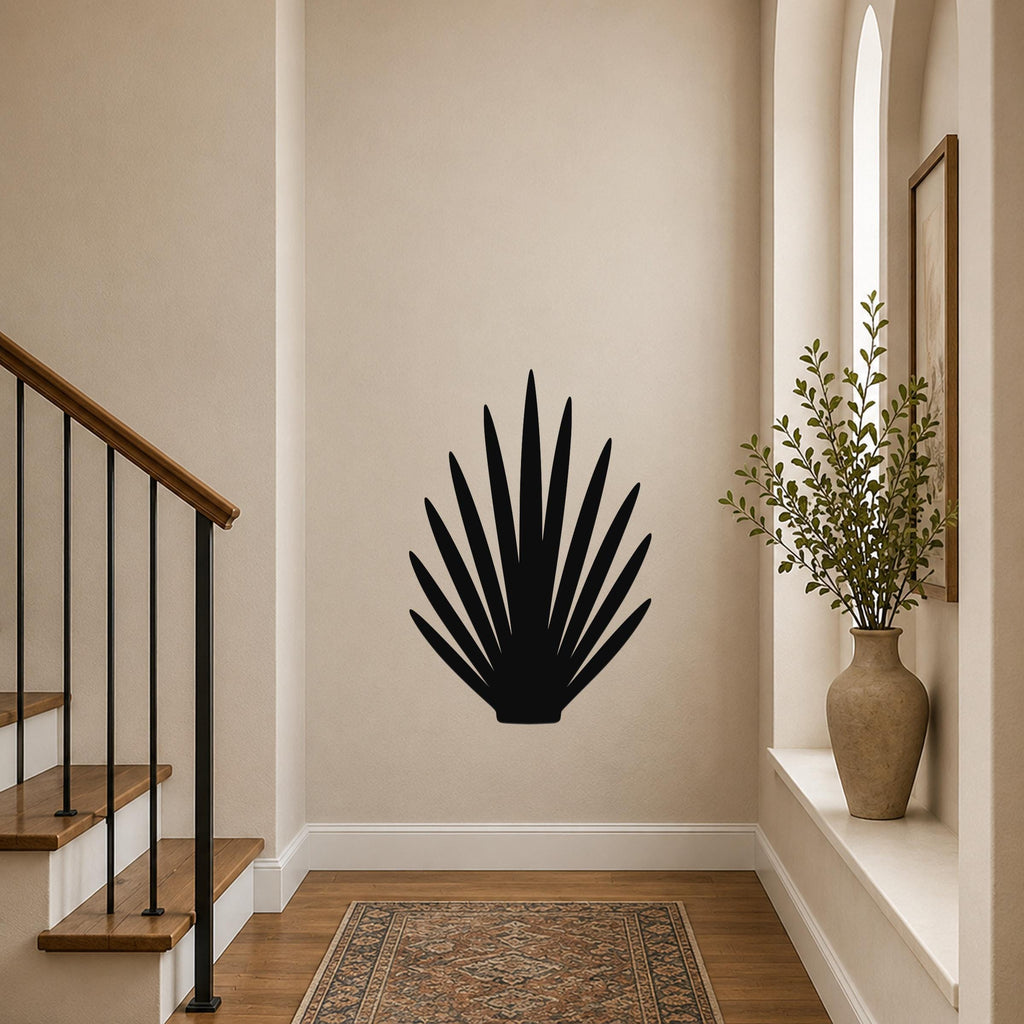 Black metal wall art in a fan-shaped grass or palm frond silhouette, mounted on a beige hallway wall near a staircase and a window with a potted plant.