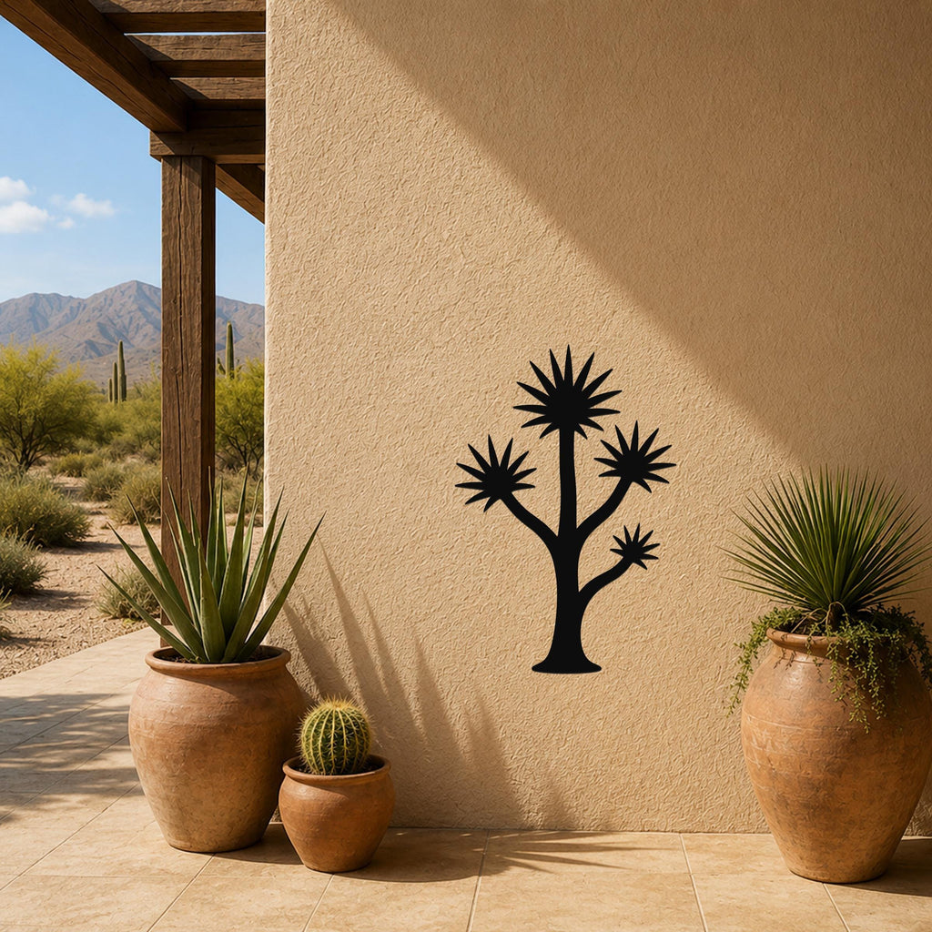 Black metal wall art silhouette of a tall yucca tree with spiky branches, mounted on a stucco outdoor wall beside potted plants and a desert landscape view.