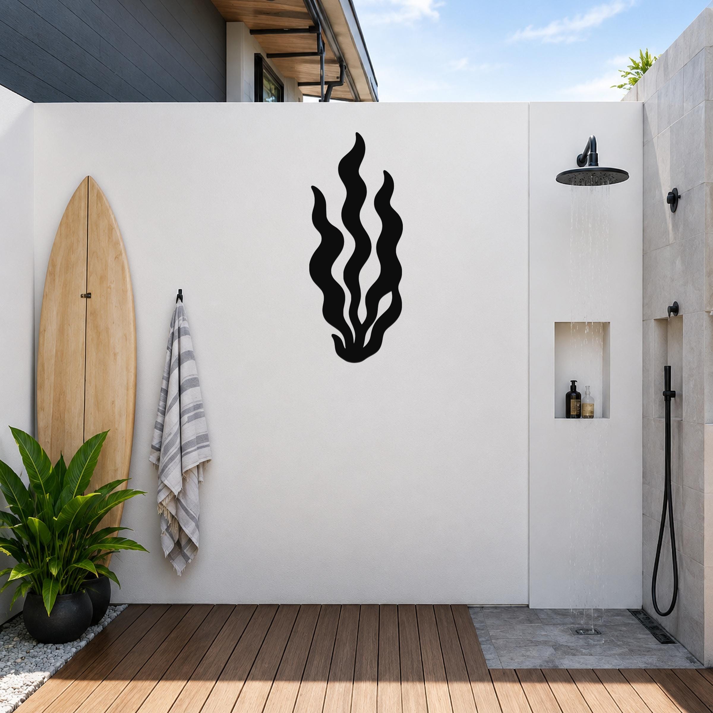 Black metal kelp wall art with flowing seaweed cutouts, mounted on a white outdoor wall above a wooden deck near a surfboard and shower.