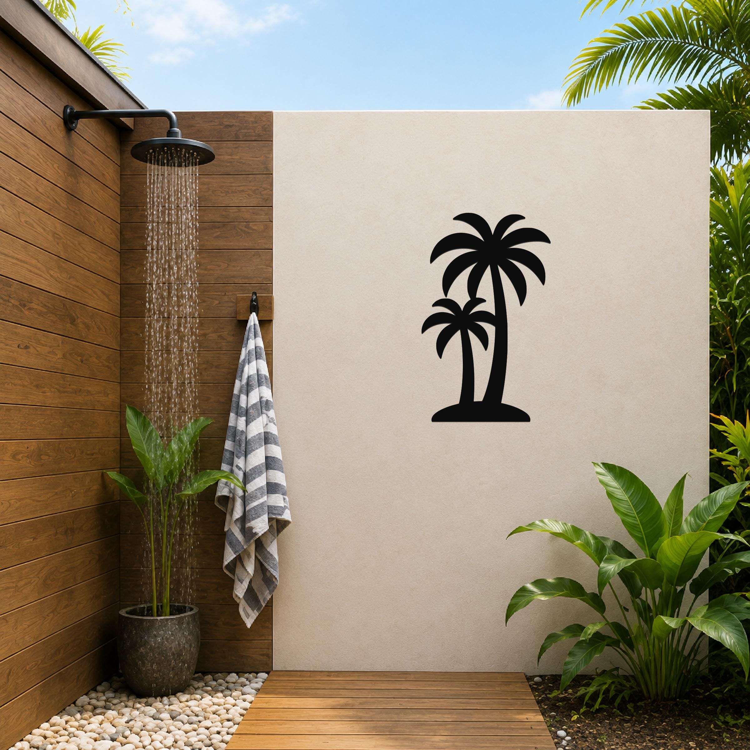 Black metal twin palm trees wall art silhouette mounted on a beige outdoor wall beside a wooden shower area with a running showerhead and tropical plants.