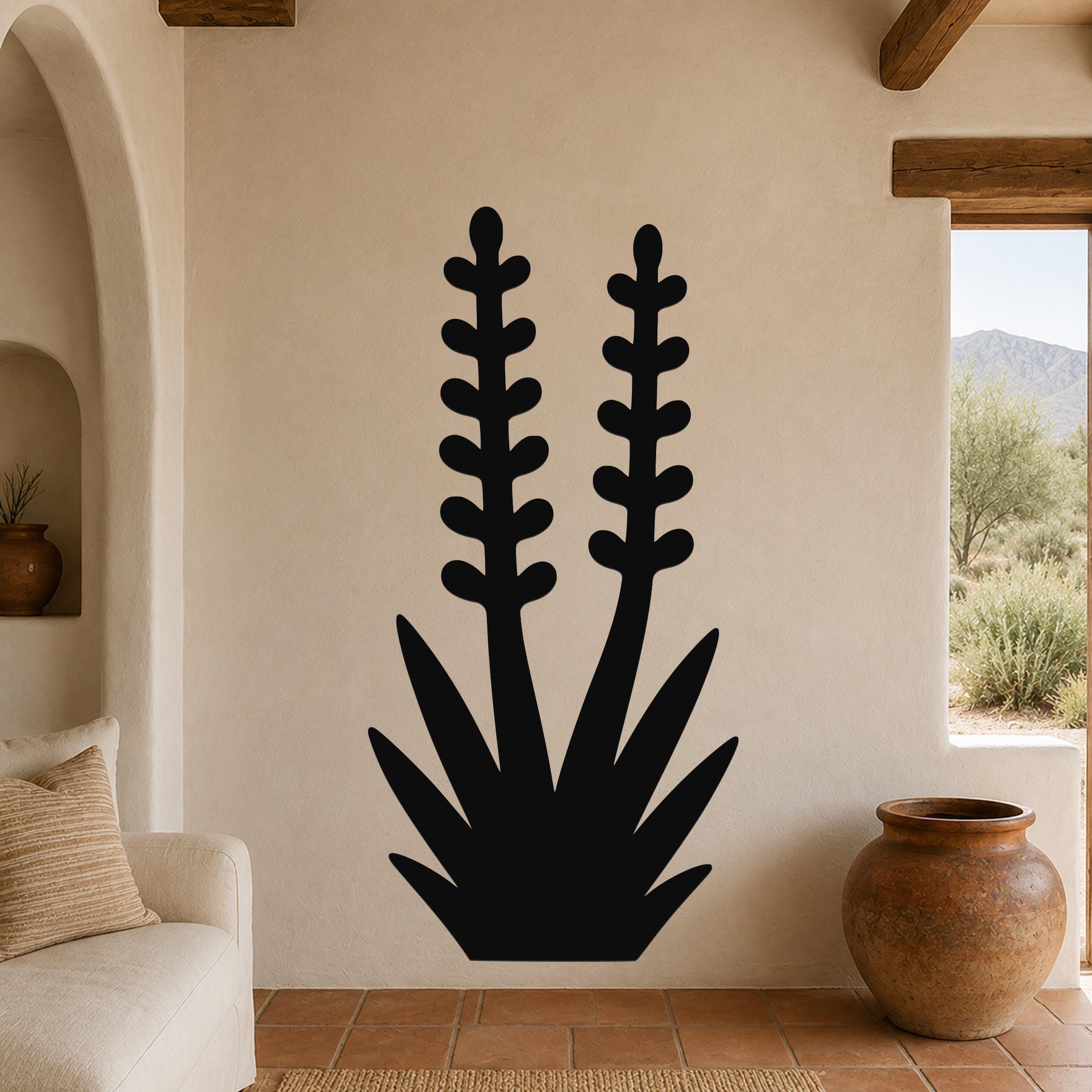Black metal wall art in a yucca plant silhouette with two tall flower stalks and spiky base, mounted on a beige wall in a living room with terracotta tile floor.