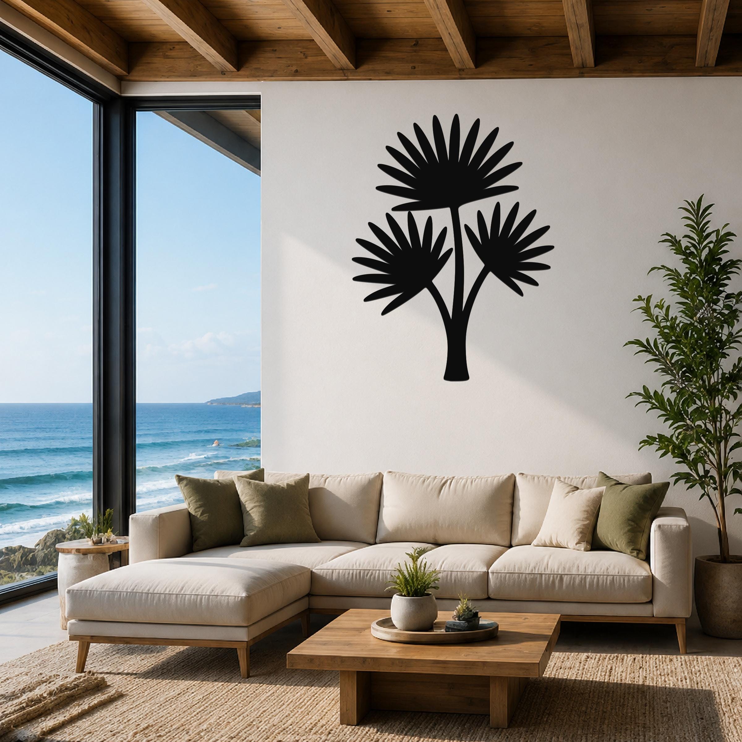 Black metal cutout palm tree wall art with three fan-shaped fronds, mounted on a white wall above a beige sectional sofa in a living room with ocean views.