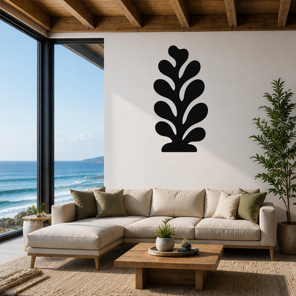 Black metal wall art in a leaf or coral silhouette, mounted on a white wall above a beige sectional sofa in a living room with ocean views.
