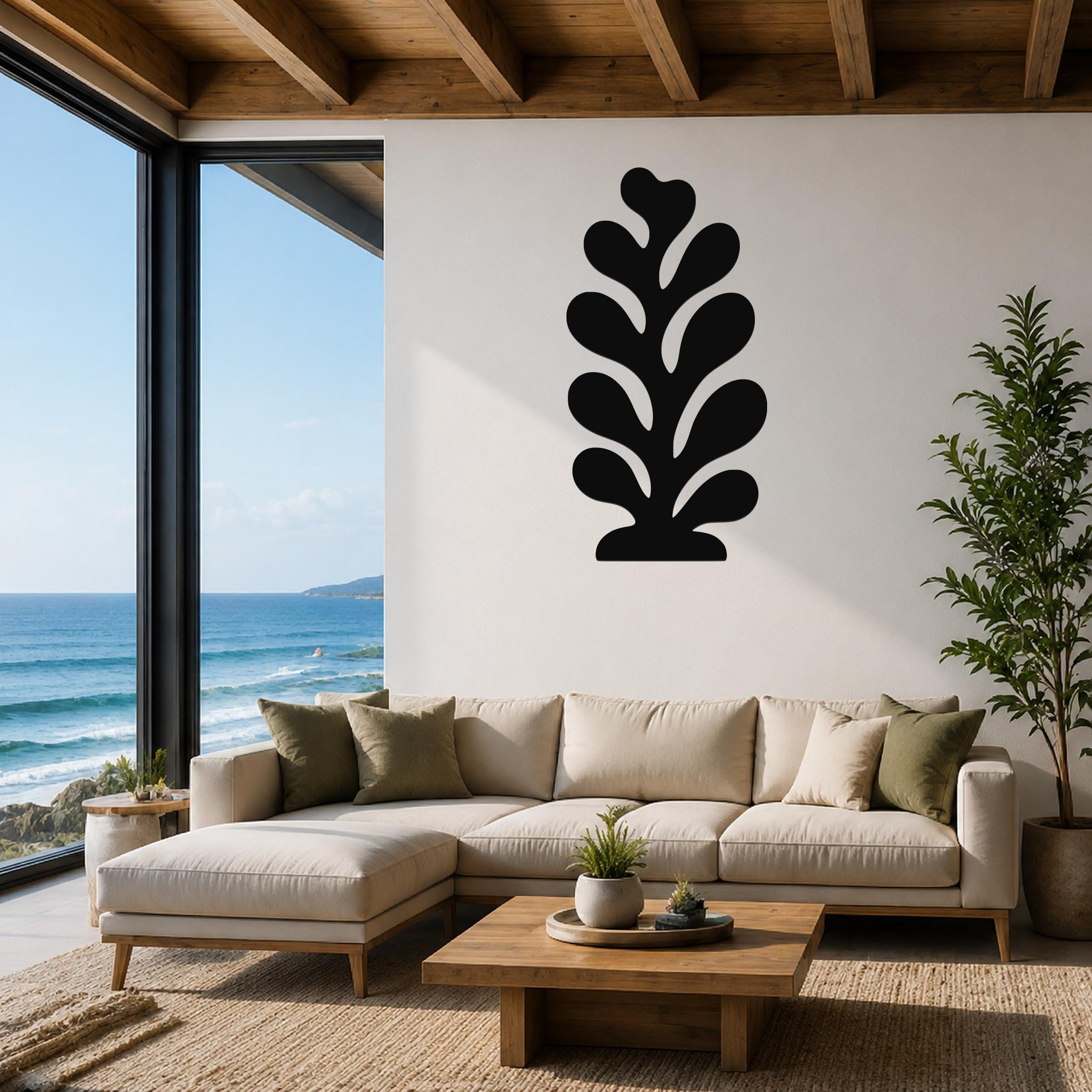 Black metal wall art in a leaf or coral silhouette, mounted on a white wall above a beige sectional sofa in a living room with ocean views.