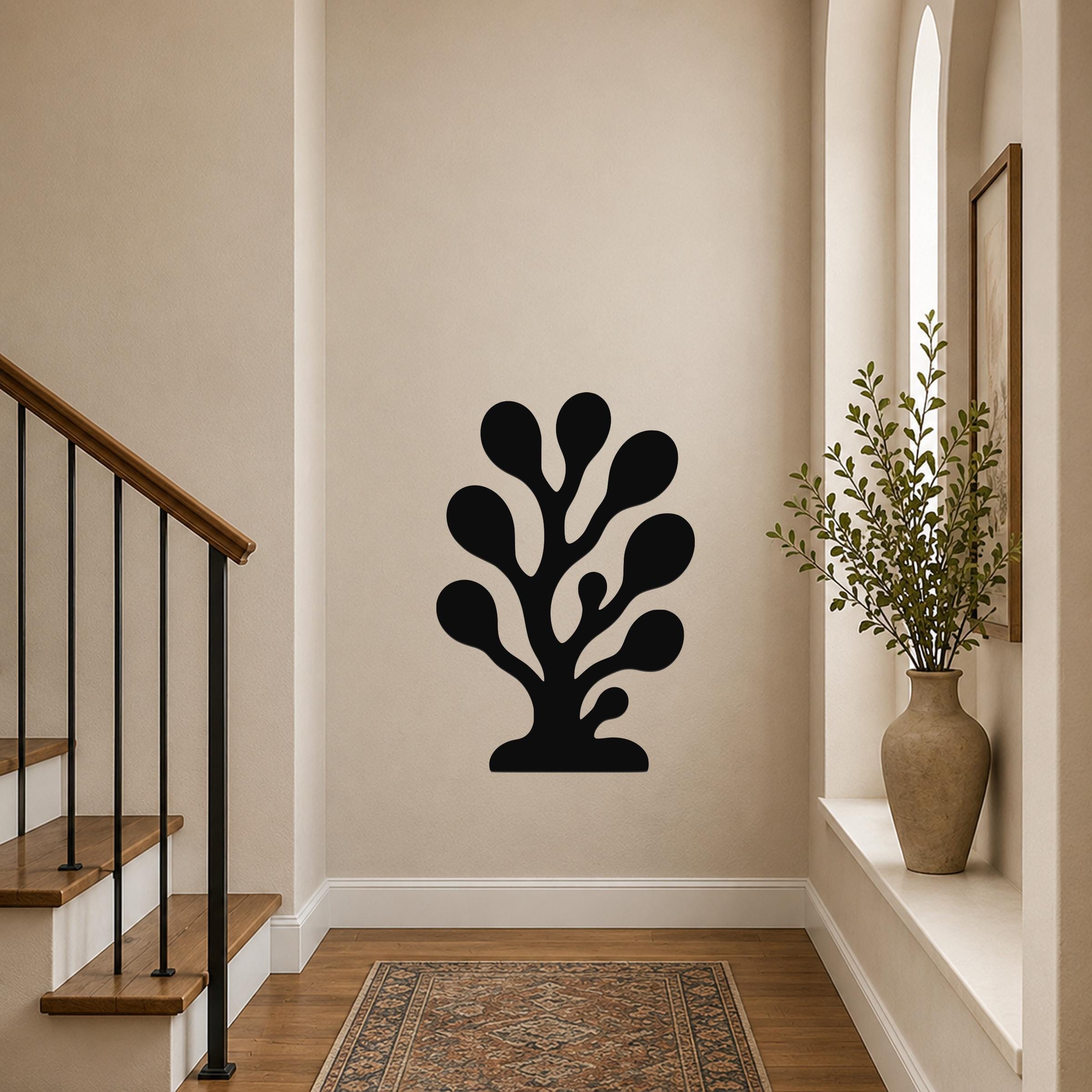 Black metal wall art in a coral or seaweed silhouette design, mounted on a beige hallway wall near a staircase and a window with a potted plant.