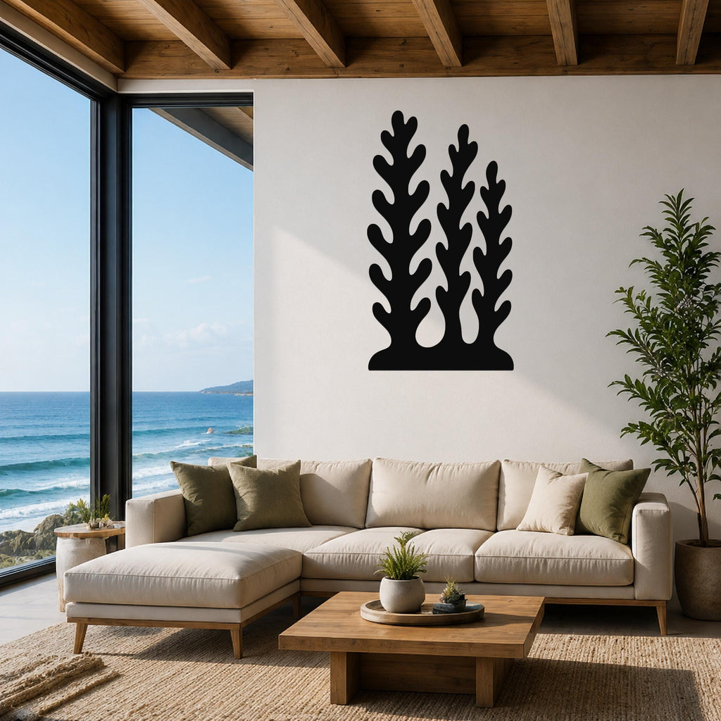 Black metal wall art with a kelp and seaweed silhouette design, mounted on a white wall above a beige sectional sofa in a living room with ocean views.