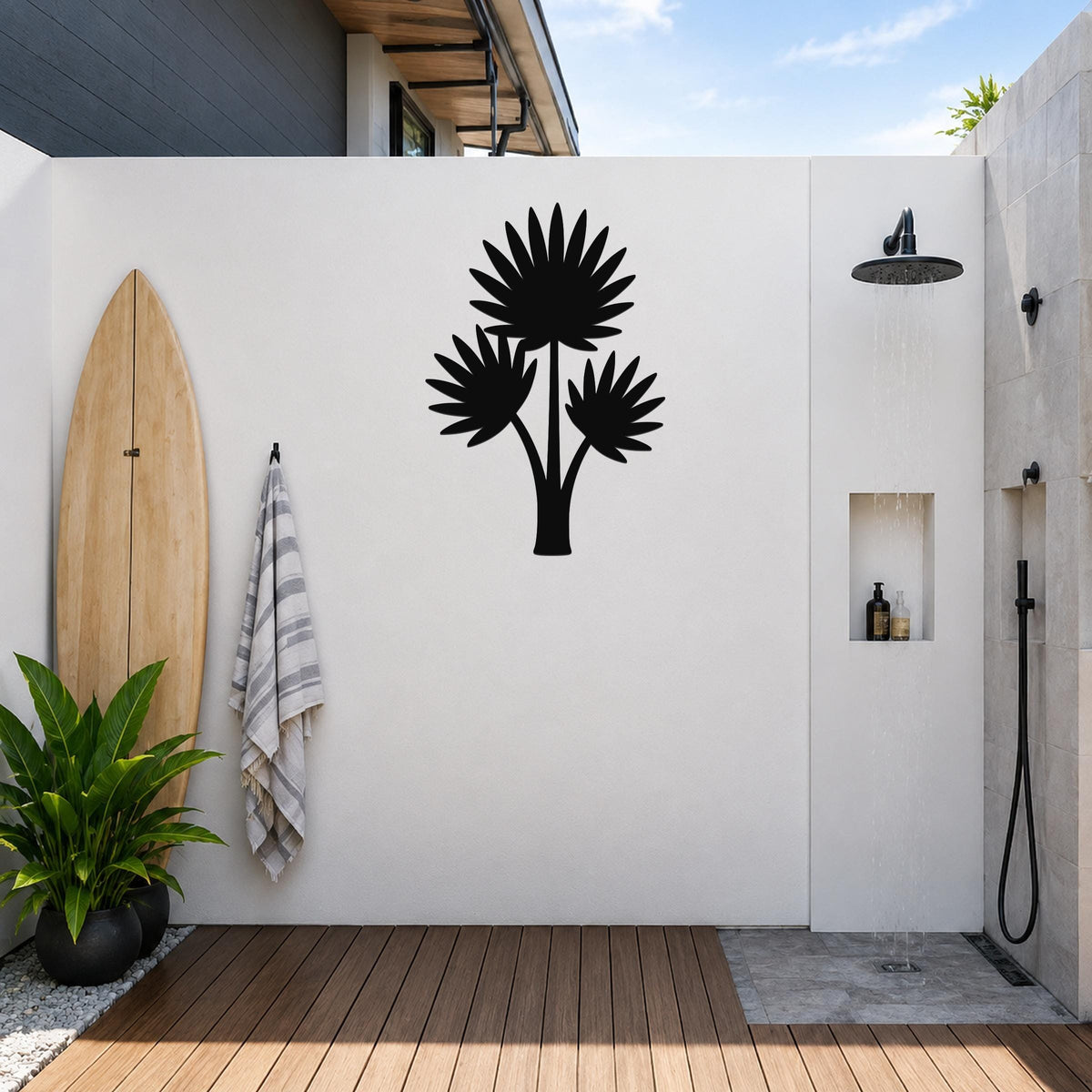 Black metal wall art of a fan palm tree silhouette, mounted on a white outdoor wall above a wooden deck near a shower area.