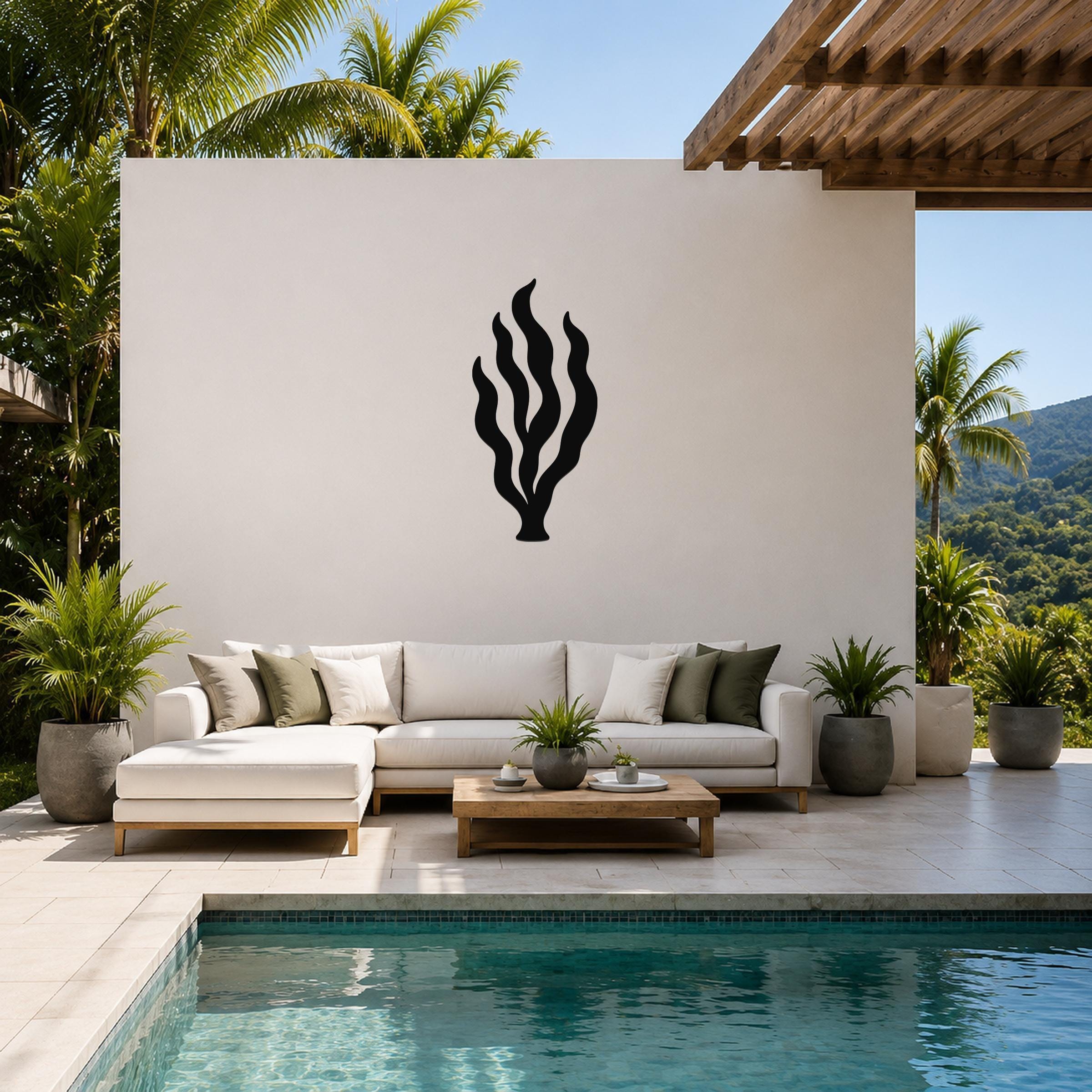 Black metal kelp seaweed wall art with flowing frond cutouts, mounted on a white outdoor wall above a white sectional sofa in a tropical patio setting.