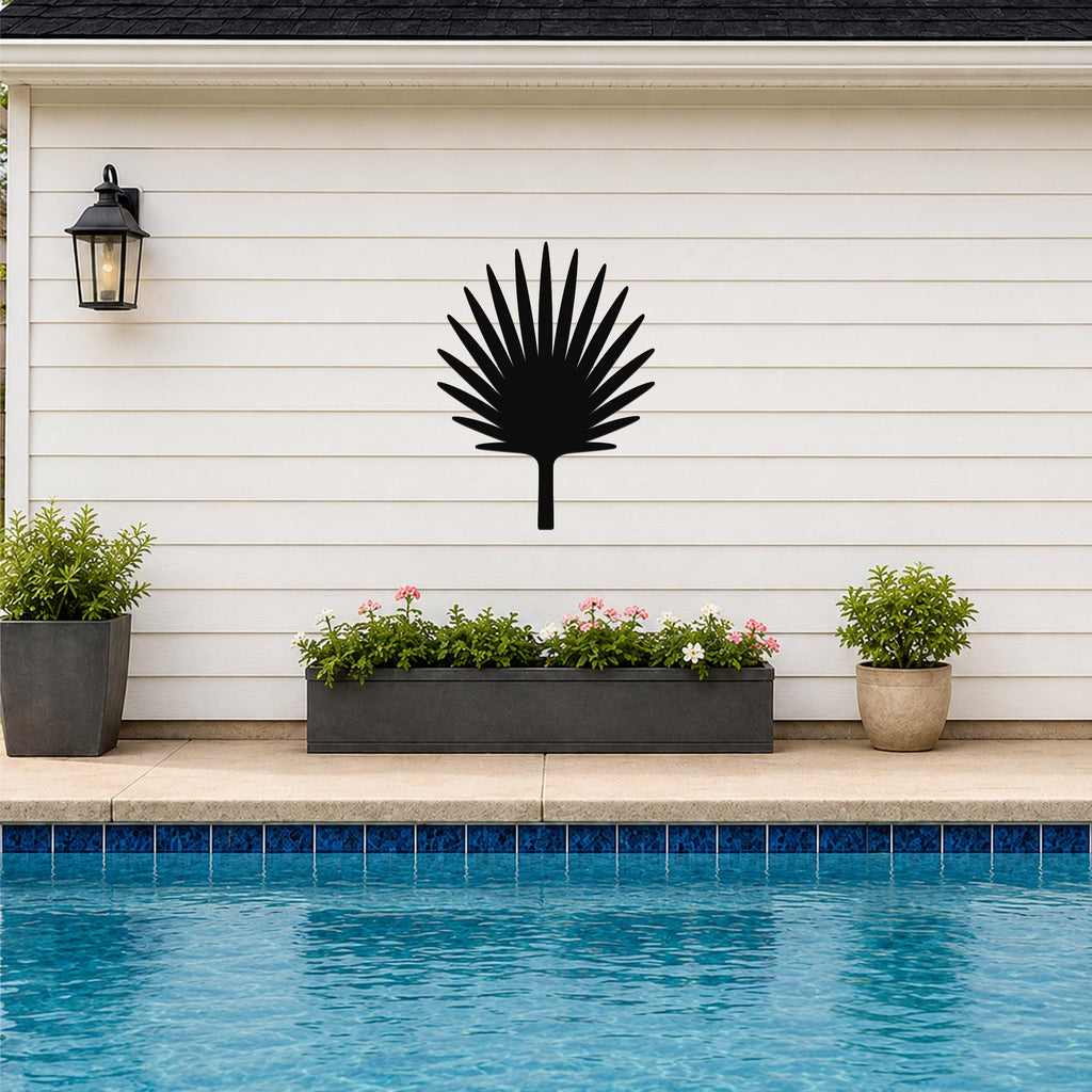 Black metal fan palm tree wall art silhouette mounted on a white horizontal siding wall above a poolside planter and patio.
