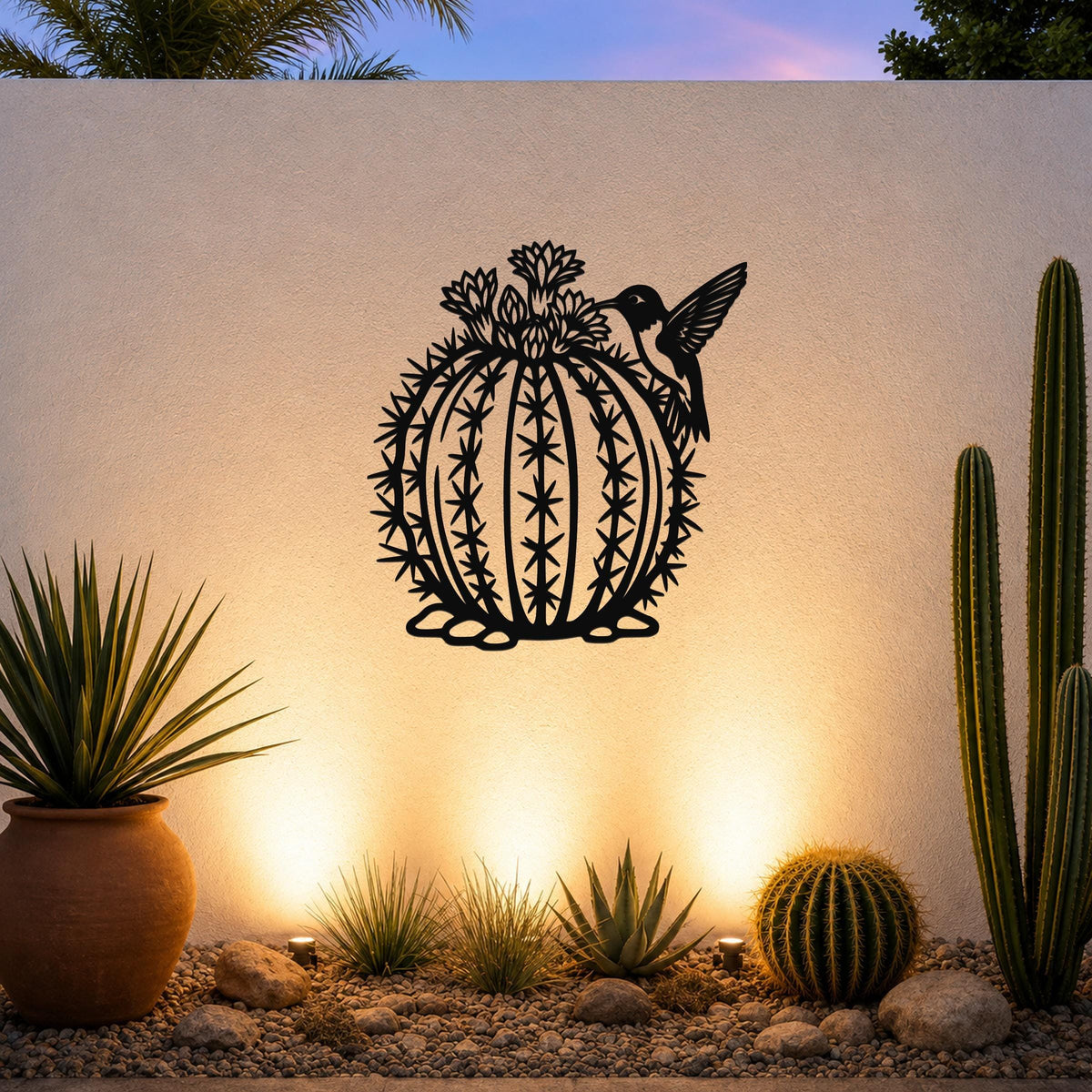 Round black metal barrel cactus wall sculpture with flower and hummingbird cutouts, mounted on a white outdoor wall above a desert garden.