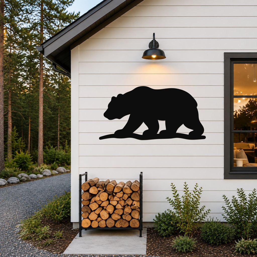 Black metal bear silhouette wall art mounted on a white horizontal siding exterior wall above a firewood rack, illuminated by a black outdoor wall light.