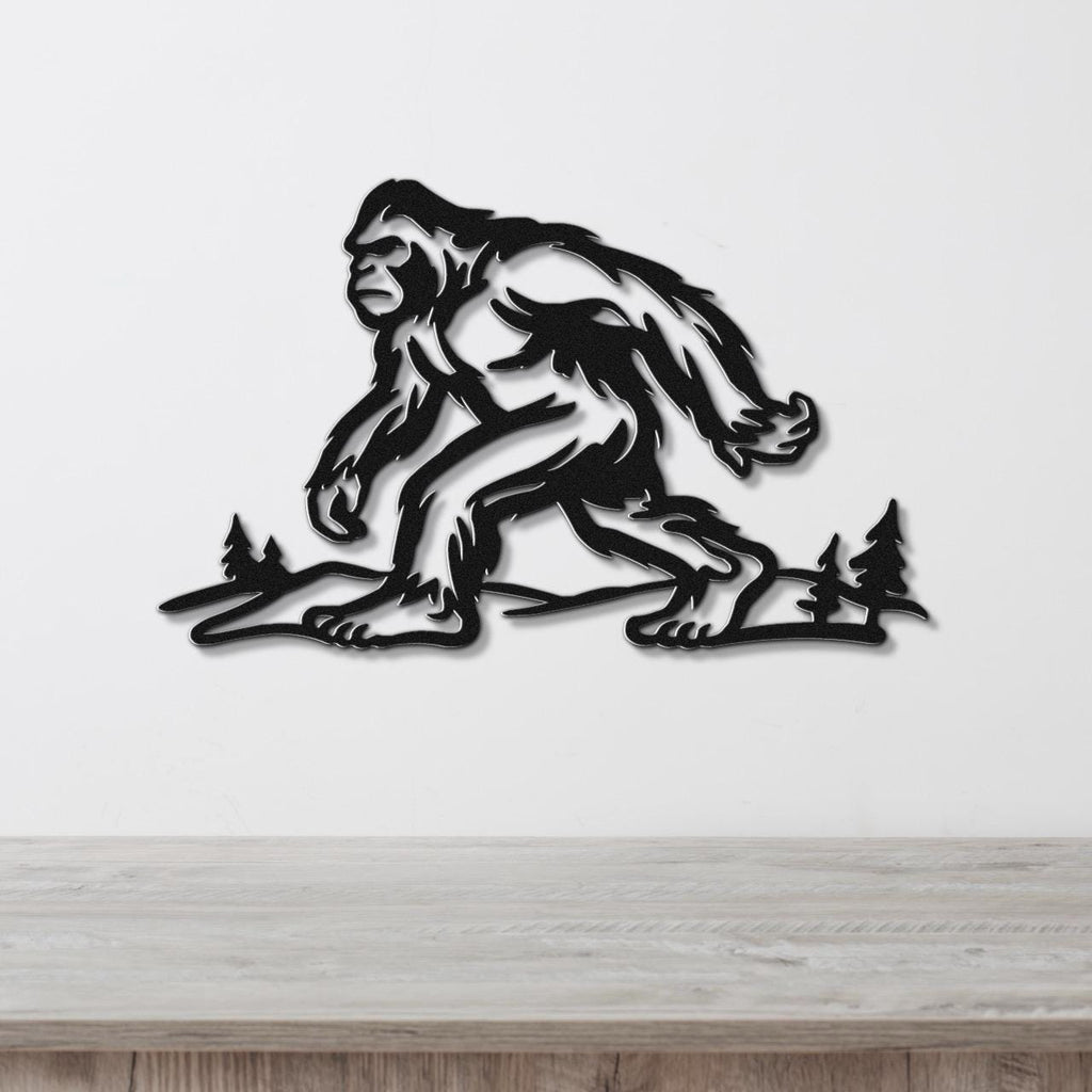 Black metal wall art silhouette of Bigfoot walking through a forest with pine trees, mounted on a white wall above a wooden surface.