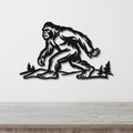 Black metal wall art silhouette of Bigfoot walking through a forest with pine trees, mounted on a white wall above a wooden surface.