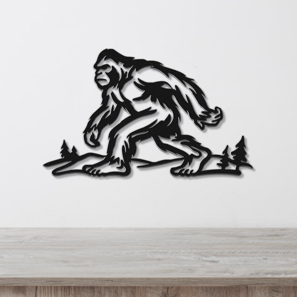 Black metal wall art silhouette of Bigfoot walking through a forest with pine trees, mounted on a white wall above a wooden surface.