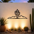 Black metal wall art silhouette of a saguaro cactus and mountain range, mounted on an outdoor wall above a desert garden with potted plants and ground lights.