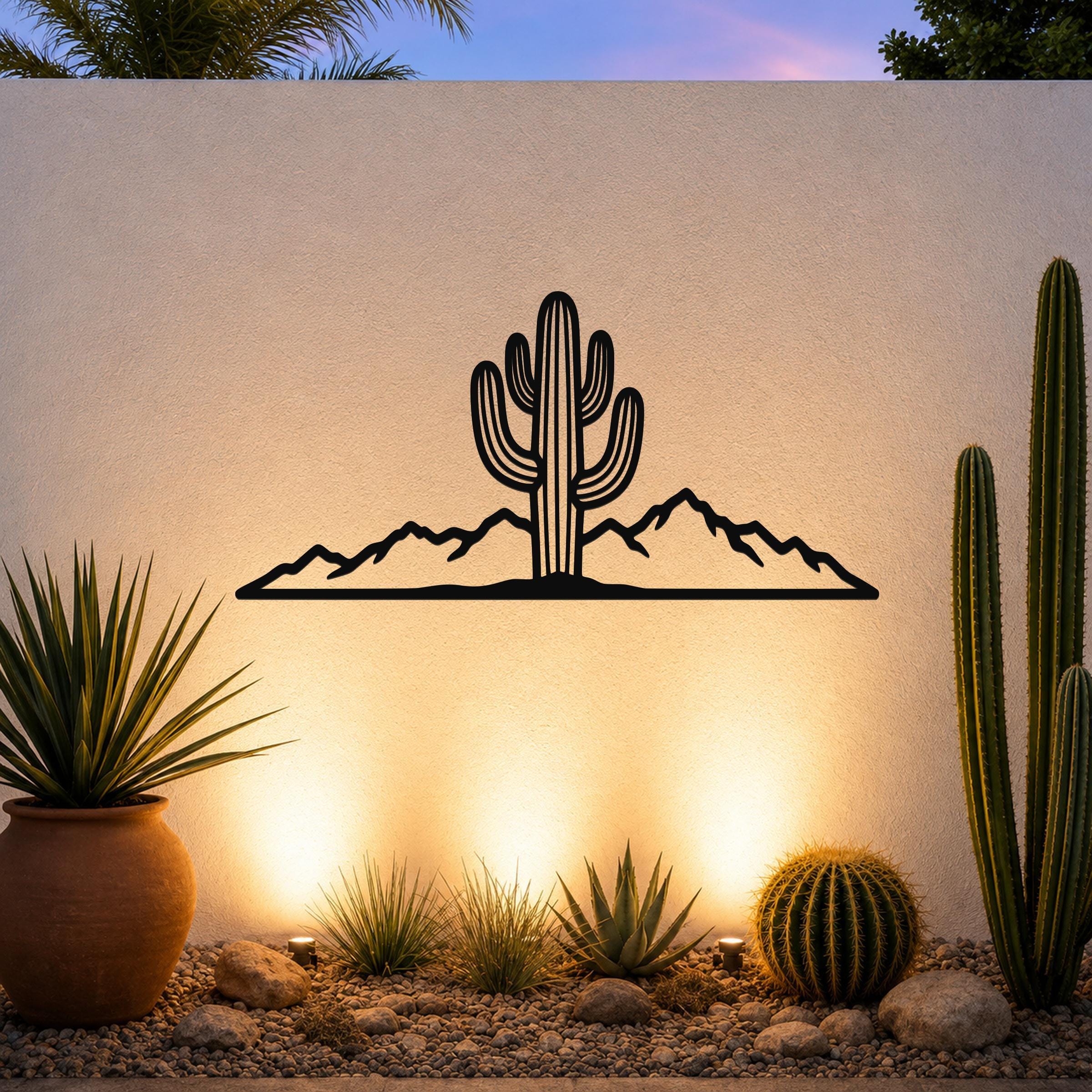 Black metal wall art silhouette of a saguaro cactus and mountain range, mounted on an outdoor wall above a desert garden with potted plants and ground lights.