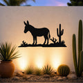 Black metal wall art silhouette of a burro and cacti, mounted on an outdoor wall above a desert garden with potted plants and ground lights.
