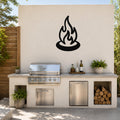Black metal campfire silhouette wall art mounted on a light-colored outdoor wall above a stainless steel grill and countertop.