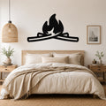 Black metal campfire wall art with flame and log silhouette, mounted above a wooden bed in a neutral bedroom.