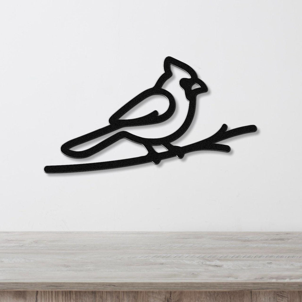 Black metal cardinal wall art in a minimalist silhouette style, perched on a branch, mounted on a white wall above a light wood surface.