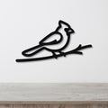 Black metal cardinal wall art in a minimalist silhouette style, perched on a branch, mounted on a white wall above a light wood surface.