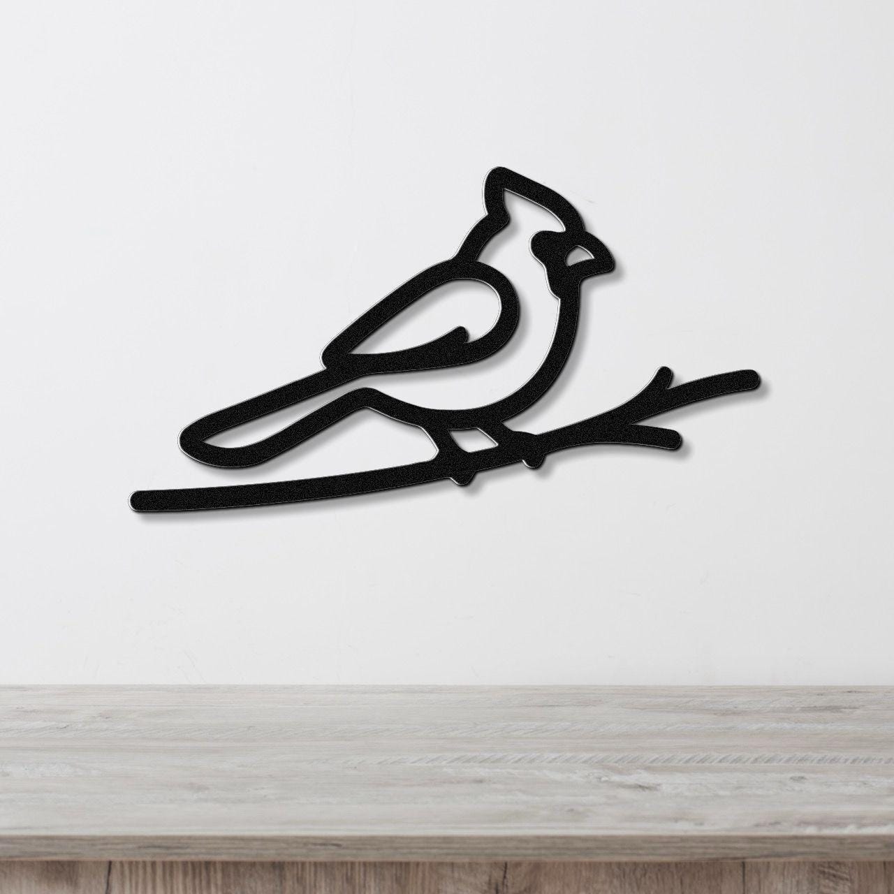 Black metal cardinal wall art in a minimalist silhouette style, perched on a branch, mounted on a white wall above a light wood surface.