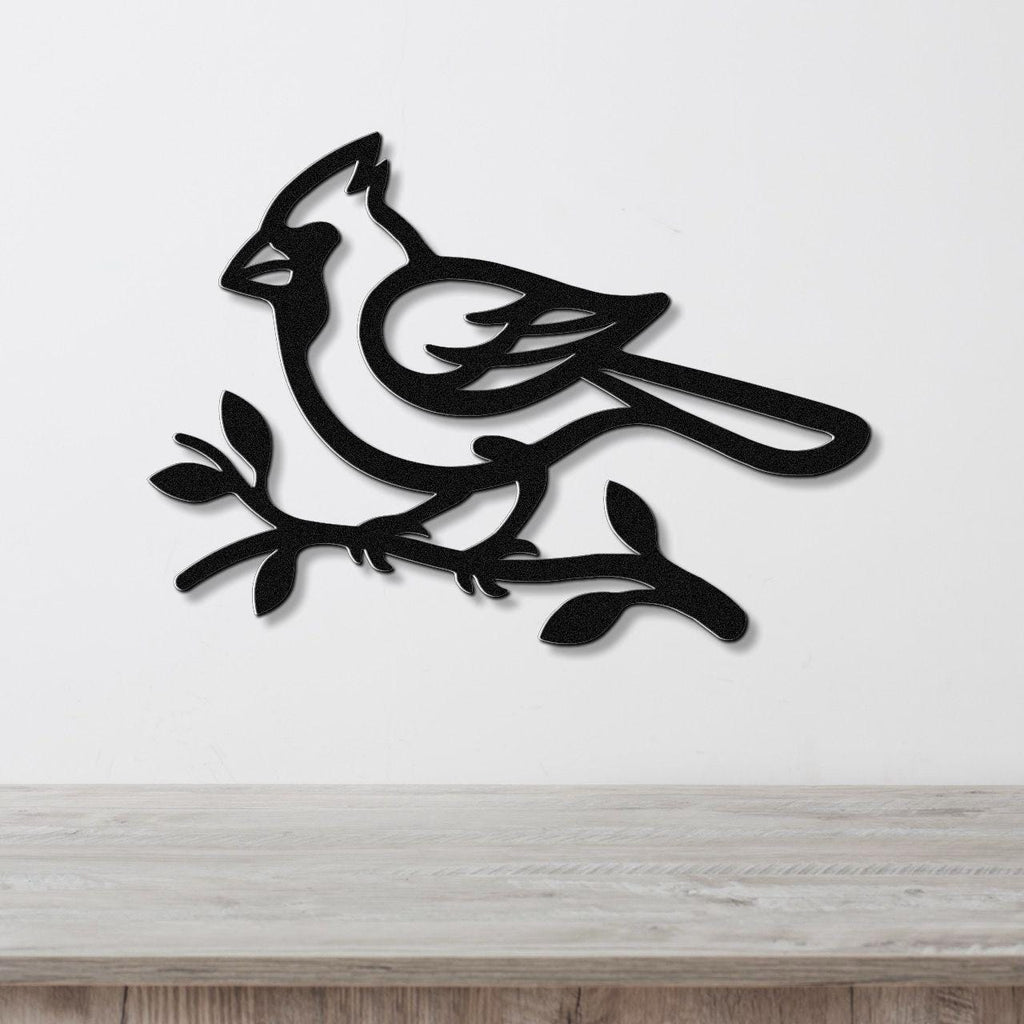 Black metal cardinal wall art in silhouette style, perched on a branch with cutout details, mounted on a white wall above a wooden surface.