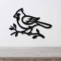 Black metal cardinal wall art in silhouette style, perched on a branch with cutout details, mounted on a white wall above a wooden surface.