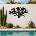 Black metal Cholla cactus wall art with branching silhouette, mounted on a white outdoor wall above a poolside garden.