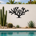 Black metal Cholla cactus wall art with branching silhouette, mounted on a white outdoor wall beside a pool and desert plants.