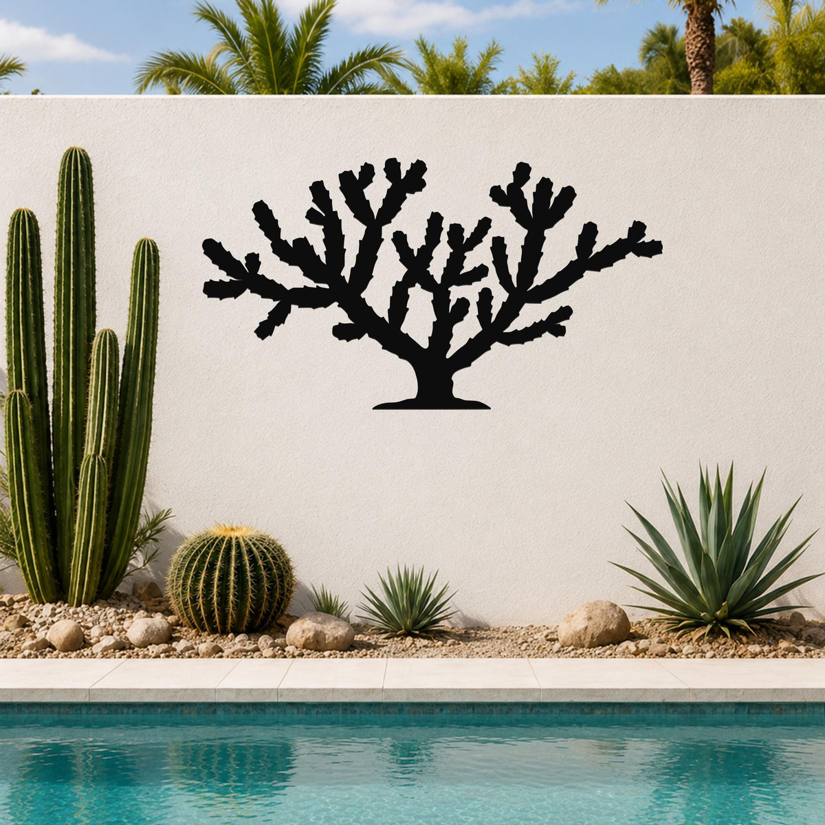 Black metal Cholla cactus wall art with branching silhouette, mounted on a white outdoor wall beside a pool and desert plants.