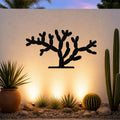 Black metal Cholla cactus wall art with branching cutouts, mounted on a textured outdoor wall above a desert garden with potted plants and ground lights.