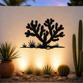 Black metal Cholla cactus wall art with branching silhouette and ground-level grass cutouts, mounted on a textured outdoor wall above a desert garden with potted succulents and ground lights.