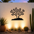 Black metal Cholla cactus wall art silhouette with branching arms and spines, mounted on a textured outdoor wall above a desert garden with potted succulents and ground lights.