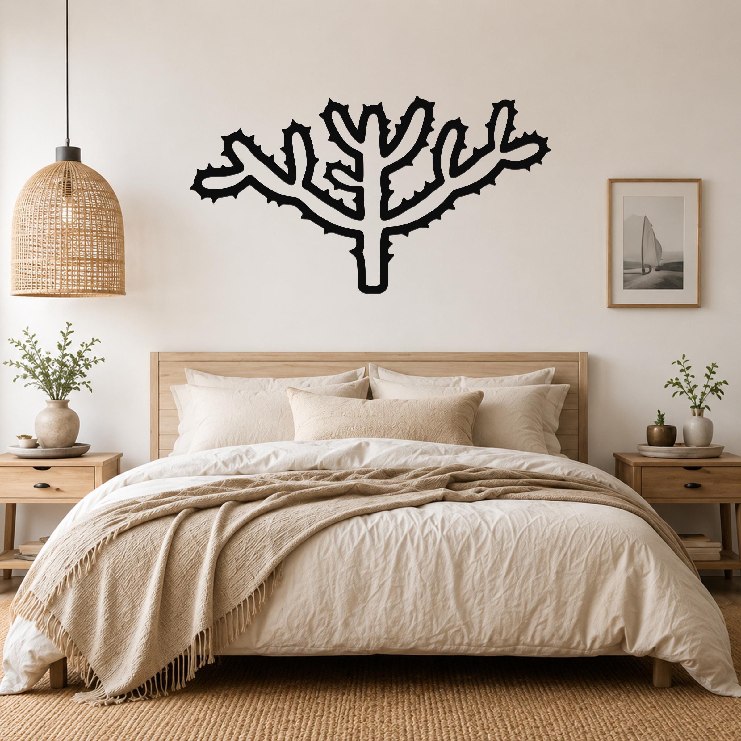 Black metal Cholla cactus wall art with spiky branch cutouts, mounted above a wooden bed in a neutral bedroom.
