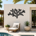 Black metal Cholla cactus wall art with intricate branch and spine cutouts, mounted on a white outdoor wall above a lounge chair near a pool.