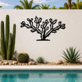 Black metal Cholla cactus wall art with detailed branch and spine cutouts, mounted on a white outdoor wall above a poolside garden.