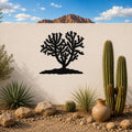 Black metal Cholla cactus wall art with detailed branch cutouts, mounted on a white outdoor wall above a desert garden with real cacti and rocks.