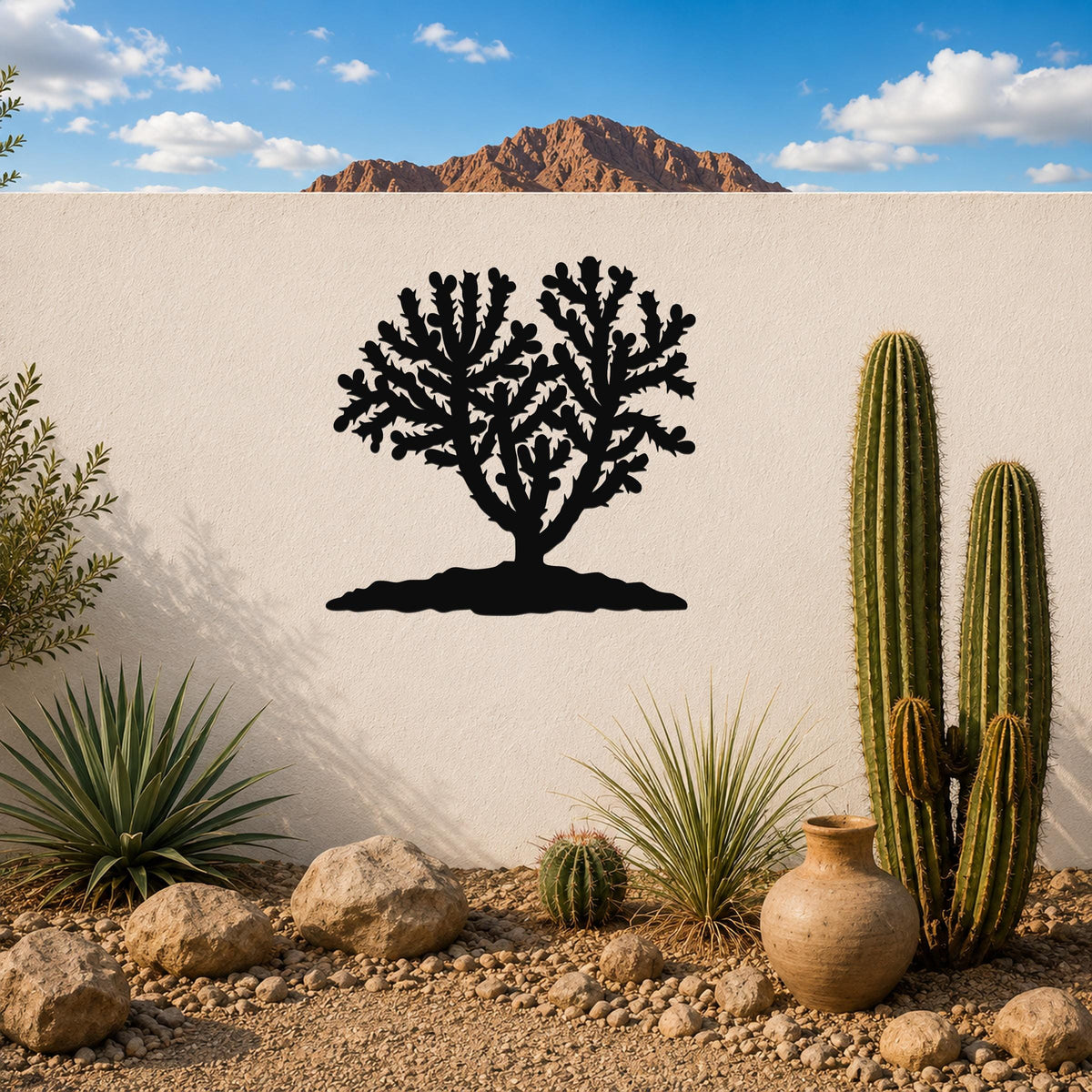 Black metal Cholla cactus wall art with detailed branch cutouts, mounted on a white outdoor wall above a desert garden with real cacti and rocks.