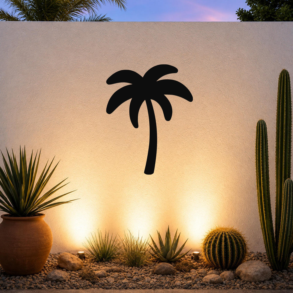 Black metal palm tree wall art silhouette mounted on a white outdoor wall above a desert garden with cacti and warm uplighting.