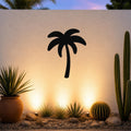Black metal palm tree wall art silhouette mounted on a white outdoor wall above a desert garden with cacti and warm uplighting.