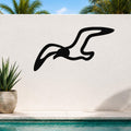 Black metal seagull wall art with flowing wing cutouts, mounted on a white outdoor wall above a pool with palm trees in the background.