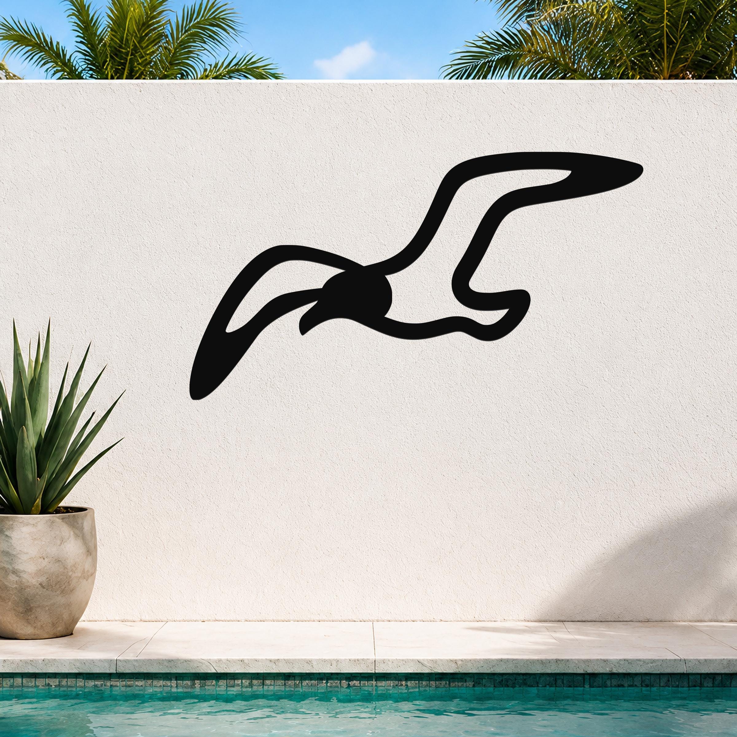 Black metal seagull wall art with flowing wing cutouts, mounted on a white outdoor wall above a pool with palm trees in the background.